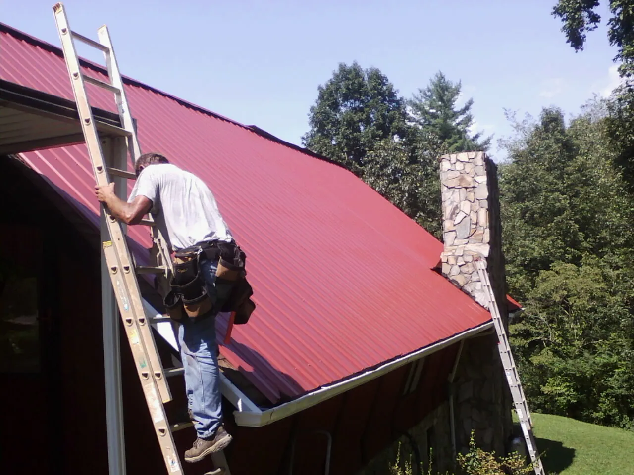 Roofer on a ladder beside a red metal roof and stone chimney — Soffit & Fascia Repair by Roofing Brothers in Harrington Park